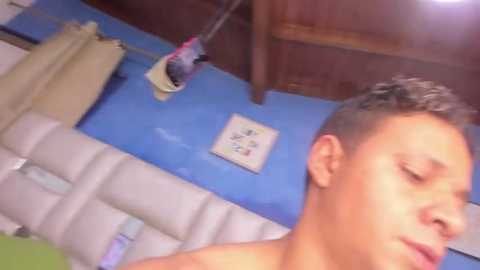 Media: A video of a young, shirtless, light-skinned man with short, dark hair, lying on a white, padded bench. The background features a blue wall, a wooden ceiling, and a hanging laundry rack.