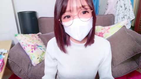 Media: Video of an Asian woman with shoulder-length dark hair, wearing glasses and a white mask, seated on a brown couch. Floral-patterned pillows and a colorful blanket are behind her in a cozy, cluttered room.