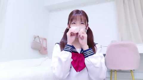 Media: Video of a young East Asian woman with long brown hair, wearing a white sailor school uniform with a red necktie, sitting indoors, applying face mask, in a modern, minimalist room with a pink chair and white walls.