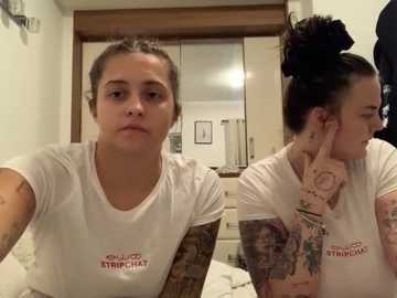 Media: Video of two tattooed women in white T-shirts, one with a brick arm tattoo, in a bedroom with a wooden headboard and framed pictures on the wall.