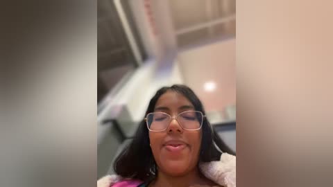 Media: A video of a young Black woman with long black hair, wearing glasses and a pink hoodie, sitting in an airplane. The background is blurred, showing the plane's interior.