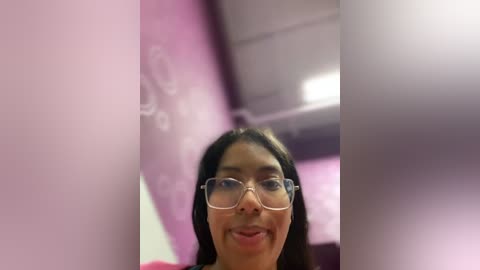 Media: Video of a smiling woman with medium skin tone, long black hair, wearing glasses, and a pink shirt, taken in a blurred, pink-tinted interior.