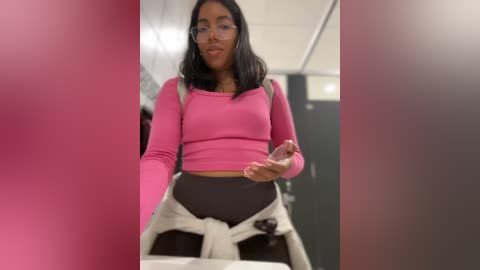 Media: Video of a young woman with medium brown skin, shoulder-length black hair, wearing a pink long-sleeve crop top, black leggings, and a white hoodie tied around her waist. She's holding a phone in a brightly lit, modern restroom.