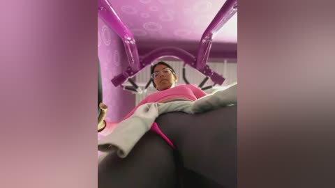 Media: Video of a young woman in a pink and white top, wearing dark leggings, sitting on a purple, circular, cushioned surface with a white ceiling.