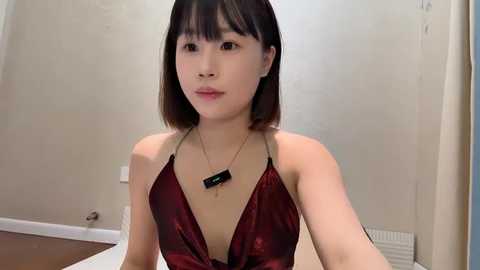 Media: Video of a young Asian woman with straight black hair, fair skin, and bangs. She wears a low-cut, shiny red halter top with a gold necklace. Background features beige walls and a radiator.