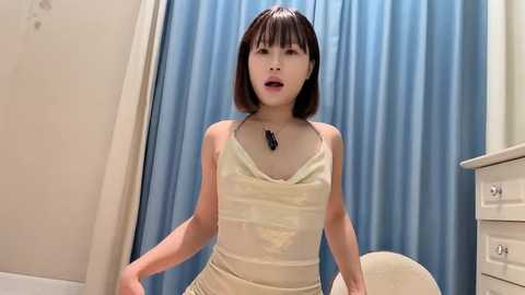 Media: Video of an Asian woman with straight black hair, wearing a yellow tank top, standing in a bathroom with blue curtains and a white dresser.