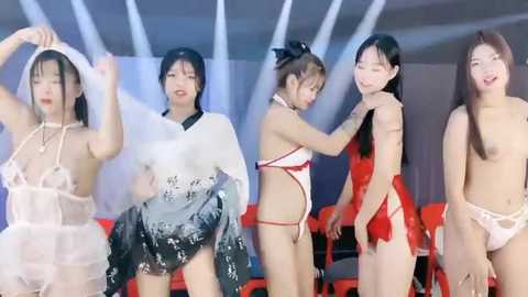 Media: Video of four Asian women in a modern room; three are partially or fully nude, one in a white kimono, another in lingerie. Background has white curtains and red chairs.