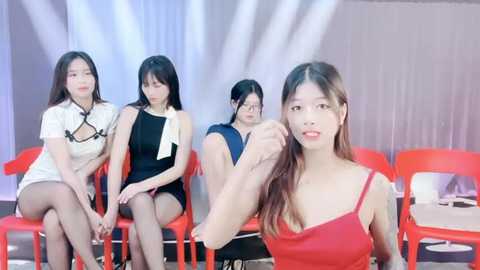 Media: Video of four Asian women in modern, casual attire seated on red chairs, one woman in red dress foreground, background with white curtains and stage lights.