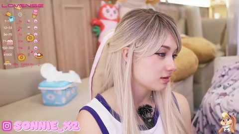 Media: Video of a blonde woman with long hair, wearing a white and purple sleeveless top, in a cozy, indoor setting with plush toys and a tissue box.