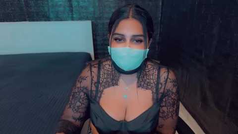 Media: Video of a woman with light brown skin and dark hair, wearing a black lace top revealing cleavage, a turquoise face mask, and intricate arm tattoos, sitting on a bed with dark bedding in a dimly lit room.