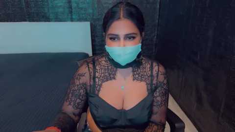 Media: Video of a young woman with medium skin tone, dark hair, and large breasts, wearing a black lace top, a blue surgical mask, and sitting in a dimly lit room with dark brick walls and a blue sofa.