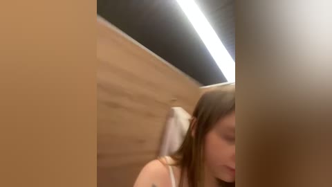 Media: Video of a young woman with long brown hair in a white headband, partially obscured by a wooden door, in a modern bathroom with a bright, white fluorescent ceiling light.