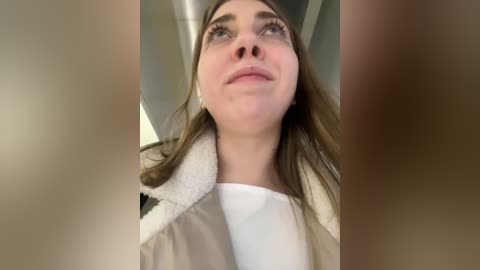 Media: A close-up video of a young woman with long, straight brown hair, wearing a white shirt and beige coat, gazing upwards, with blurry hands in the foreground.