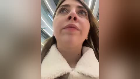 Media: A video of a young woman with fair skin and light brown hair, wearing a brown coat with a white fur collar, gazing upward in a modern, well-lit indoor setting with glass panels and soft lighting.