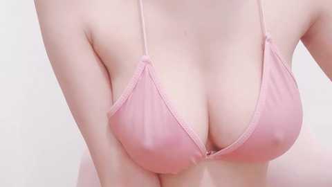 Media: Video of a fair-skinned woman with large breasts, wearing a sheer, pink, triangle bikini top, revealing nipples. She has a smooth, light complexion and the background is a plain, light beige.