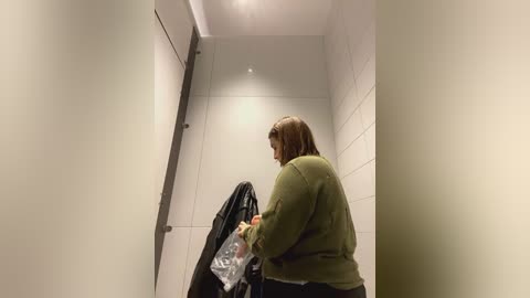 Media: Video of a woman in a bathroom stall with white walls, wearing a green sweater and holding a plastic bag and a black jacket.