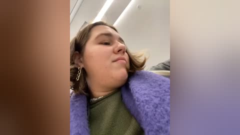 Media: Video of a young woman with shoulder-length brown hair, wearing a green sweater, leaning back in a plane seat. A purple blanket covers her legs. The background shows airplane cabin interior with white walls and overhead lights.