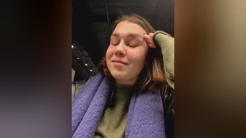 Media: Video of a young woman with fair skin, light brown hair, and closed eyes, wearing a green sweater, leaning back in a car seat with a purple blanket over her lap.