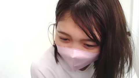 Media: A video of an East Asian woman with straight, shoulder-length dark hair, wearing a white mask, looking down, in a soft pink top, against a plain white background.