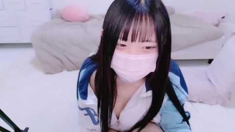 Media: Video of an East Asian woman with long black hair, wearing a pink mask, a blue and white jacket, and a low-cut white top, kneeling on a white floor, in a minimalist room with a white bed and soft pink pillow.