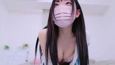 Media: A video of an East Asian woman with long black hair, wearing a pink face mask and a revealing white top, in a minimalist white room.