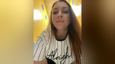 Media: Video of a young Caucasian woman with long, straight brown hair, wearing a white pinstriped shirt with \"Wilshire\" written in black script, against a yellow and blue patterned background.