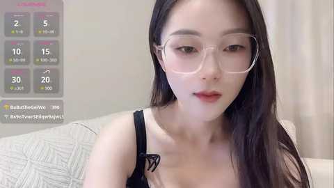 Media: A video of an Asian woman with fair skin, long black hair, and large, clear glasses, wearing a black bra, sitting on a white couch. Background shows a digital calendar with days marked.