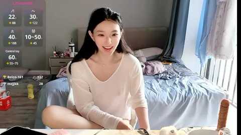 Media: Video of a young East Asian woman with long black hair, wearing a white sweater, sitting on a bed in a messy bedroom with a blue bedspread and various items on the floor.