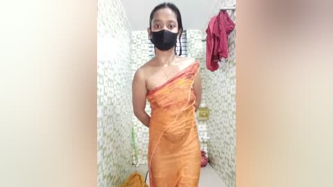 Media: Video of a woman in an orange sari, black face mask, and dark hair, standing in a tiled shower with white and green mosaic tiles, and a red towel hanging.