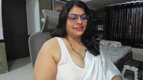 Media: Video of a middle-aged South Asian woman with dark hair, wearing glasses, a white top, and a gold necklace, seated in a modern bedroom with a gray chair, floral-patterned bedspread, and dark curtains.