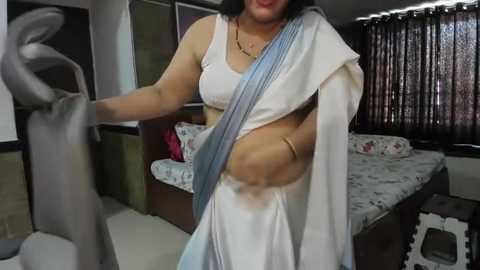 Media: Video of a South Asian woman in a white sari, adjusting a statue in a modern bedroom with a bed and patterned curtains.