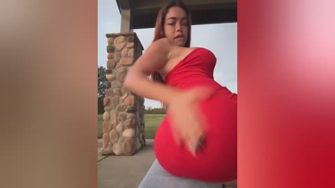 Media: Video of a curvy, light-skinned Latina woman in a tight, red dress, sitting on a stone pillar outside, with a blurred background of greenery and a wooden roof.