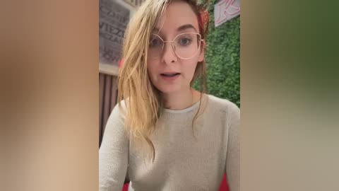 Media: Video of a fair-skinned, blonde-haired woman with glasses, wearing a light grey sweater, standing in a restaurant with green walls and a pizza menu visible.