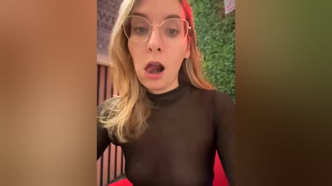 Media: A video of a young woman with fair skin and blonde hair, wearing glasses and a sheer black top, captured from a close-up angle. She has an open-mouthed expression.