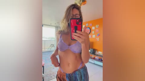 Media: Video of a blonde woman with glasses in a light purple lace bra and plaid pants, taking a selfie in a bright, orange-painted room with a pool view.