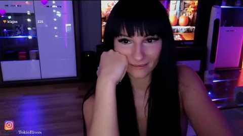 Media: Video of a young woman with long black hair, wearing a black tank top, leaning on her elbow, looking contemplative. Background includes a modern kitchen with white appliances, and a TV displaying food images.