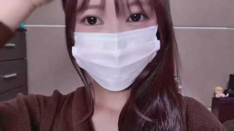 Media: A video of an East Asian woman with long, straight brown hair, wearing a white surgical mask, a brown cardigan, and light makeup. She's indoors, with a dresser and a small toy in the background.