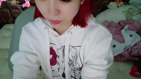 Media: Video of a young, fair-skinned woman with bright red hair, wearing a white hoodie with a cartoon character graphic, lying on a bed with pink sheets and scattered clothes in the background.