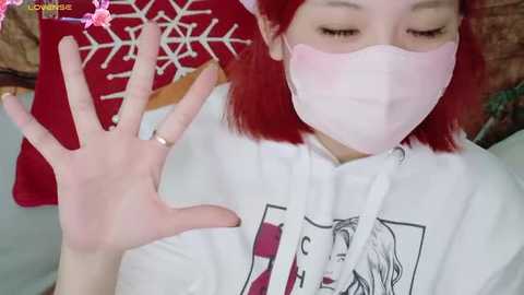 Media: Video of an Asian woman with red hair, wearing a pink face mask and white hoodie featuring anime characters. She waves her hand, set against a red blanket with a white snowflake pattern.