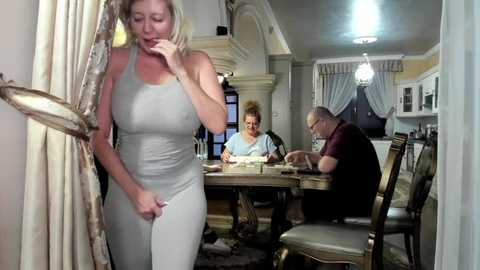 Media: A video of a woman in a tight grey dress, standing in a doorway, looking upset. A man and woman sit at a dining table in a dimly lit room with white curtains.