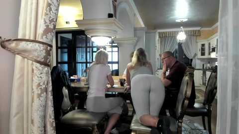 Media: Video of three people in a dimly lit, ornate room with a chandelier, cream-colored curtains, and a large window. Two women in tight leggings and a man in a suit are seated at a wooden table.