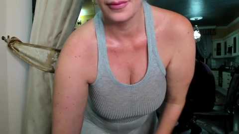 Media: A video of a middle-aged, fair-skinned woman with light brown hair, wearing a light gray, ribbed tank top that highlights her large breasts, standing in a dimly lit kitchen with beige curtains and a chandelier.