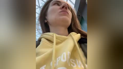 Media: Video of a young woman with long brown hair, fair skin, wearing a yellow hoodie with white text, looking up with a serene expression. Background shows a modern, glass-walled interior.