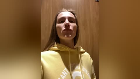 Media: Video of a young woman with long, straight brown hair, wearing a yellow hoodie with white text. She stands in a dimly lit, beige-walled room with a wooden door in the background.