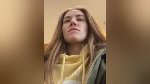 Media: Video of a young Caucasian woman with long, light brown hair, wearing a yellow hoodie, standing in a blurred, beige indoor setting.