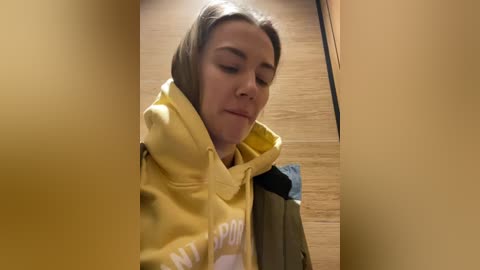 Media: Video of a young Caucasian woman with light skin, light brown hair, and a yellow hoodie, standing in a dimly lit, narrow hallway with wooden floors, looking down.