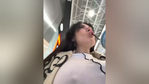 Media: Video of a young woman with fair skin and long dark hair, wearing a white top with black designs, sticking out her tongue in a playful manner. Background features a modern indoor setting with a white ceiling and geometric patterns.