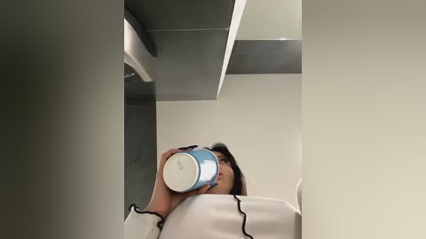 Media: A video of a woman with shoulder-length dark hair, wearing a white jacket, drinking from a blue and white thermos in a modern, minimalist office space with grey walls and sleek lighting.