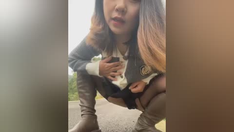 Media: Video of an Asian woman with long hair, squatting, wearing a gray sweater, black skirt, and thigh-high boots, in a park.