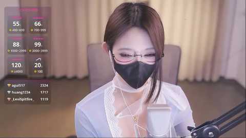 Media: Video of an Asian woman with glasses and a black face mask, wearing a white lace blouse, seated at a desk with a microphone. Background shows beige curtains and a wooden floor.